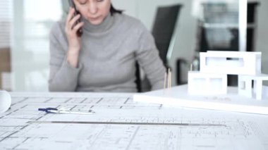 Mimar kadın telefonda konuşuyor, ayrıntılı planları ve modern ev modelini inceliyor, ofis çalışma alanında tasarım, planlama ve profesyonel iletişime odaklanıyor,