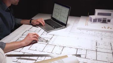 Mimar, pusula kullanarak büyük bina planlarına hassas ölçümler çiziyor, çağdaş bir ev tasarımı için mimari planlar üzerinde çalışıyor, bir dizüstü bilgisayar ve fiziksel model ile.