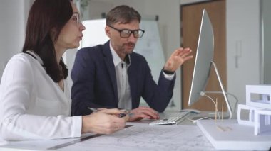 Mimarlar, modern ofis ortamında tasarım ayrıntıları ve inşaat planlarını tartışan planlar ve ölçekli bir model üzerinde işbirliği yapıyorlar. İş ve mimari kavramı