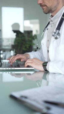 Laboratuvar önlüğü ve steteskop giyen bir doktor cam bir masadaki modern bir dizüstü bilgisayarda yazı yazıyor, sağlık bilgilerini yönetiyor. Tıp, sağlık ve bilim konsepti