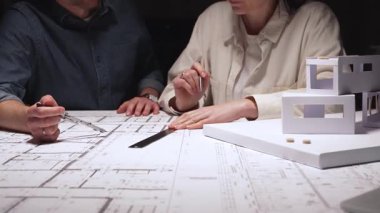 Mimari planlar ve kağıt ev modeli üzerinde yoğunlaşan profesyoneller, gelecekte bir konut veya ticari kalkınma için inşaat unsurlarını dikkatle ölçüyor ve tartışıyor