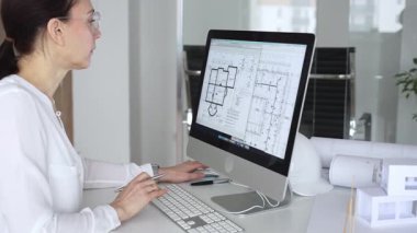 Mimari planlar ve bina modelleriyle çevrili bir masada, bir masaüstü bilgisayarı ve klavyeyi kullanarak gözlük takan mimar. İş ve mimari kavramı