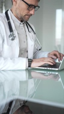 Laboratuvar önlüğü ve steteskop giyen bir doktor cam bir masadaki modern bir dizüstü bilgisayarda yazı yazıyor, sağlık bilgilerini yönetiyor. Tıp, sağlık ve bilim konsepti
