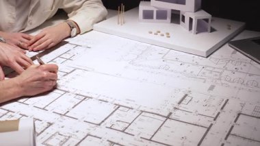 Mimarlar planlar ve ölçek modeli, cetvel ve kalemle planları ölçme ve işaretleme konusunda işbirliği yaparken, modern bir ofis iş yerindeki dizüstü bilgisayar ve evraklara atıfta bulunuyorlar.