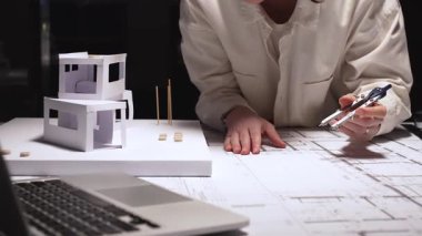 Profesyonel mimar, masa üzerinde pusula ve ölçekli ayrıntılı planlar hazırlıyor, modern ev inşaatını hassasiyetle ve yaratıcılıkla planlarken konut modelini gözden geçiriyor.