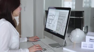 Mimari planlar ve bina modelleriyle çevrili bir masada, bir masaüstü bilgisayarı ve klavyeyi kullanarak gözlük takan mimar. İş ve mimari kavramı