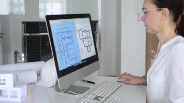 Mimari planlar ve bina modelleriyle çevrili bir masada, bir masaüstü bilgisayarı ve klavyeyi kullanarak gözlük takan mimar. İş ve mimari kavramı