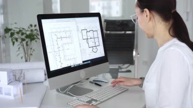 Mimari planlar ve bina modelleriyle çevrili bir masada, bir masaüstü bilgisayarı ve klavyeyi kullanarak gözlük takan mimar. İş ve mimari kavramı