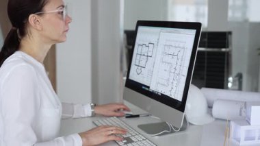 Mimari planlar ve bina modelleriyle çevrili bir masada, bir masaüstü bilgisayarı ve klavyeyi kullanarak gözlük takan mimar. İş ve mimari kavramı