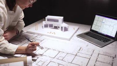 Mimar, bir masa üzerine dikkatlice bina planları çizer, bir dizüstü bilgisayar ve konut ve yapı için fiziksel bir model kullanarak modern bir tasarım yaratır.
