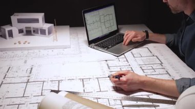 Mimar, pusula kullanarak büyük bina planlarına hassas ölçümler çiziyor, çağdaş bir ev tasarımı için mimari planlar üzerinde çalışıyor, bir dizüstü bilgisayar ve fiziksel model ile.