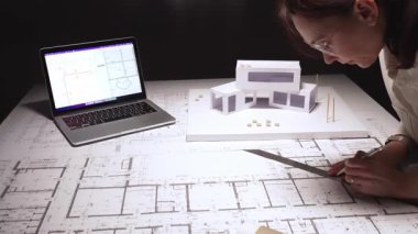 Mimar, bir masa üzerine dikkatlice bina planları çizer, bir dizüstü bilgisayar ve konut ve yapı için fiziksel bir model kullanarak modern bir tasarım yaratır.