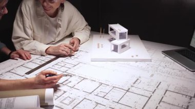 Mimarlar planlar ve ölçek modeli, cetvel ve kalemle planları ölçme ve işaretleme konusunda işbirliği yaparken, modern bir ofis iş yerindeki dizüstü bilgisayar ve evraklara atıfta bulunuyorlar.