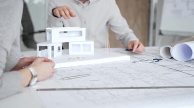 Bir konut projesinde işbirliği yapan mimarlar, planlar ve ölçekli bir modeli bir masada inceleyerek, tasarım detaylarını, inşaat planlarını ve gelecekteki gelişimi tartışarak,