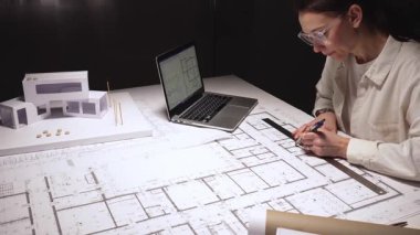 Kadın mimar konut planları tasarlıyor, cetvel ve kalemle planlar hazırlıyor, dizüstü bilgisayar teknolojisi ve mühendislik araçları kullanarak modern ev yapımı için bir model geliştiriyor.