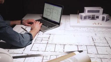 Mimar, pusula kullanarak büyük bina planlarına hassas ölçümler çiziyor, çağdaş bir ev tasarımı için mimari planlar üzerinde çalışıyor, bir dizüstü bilgisayar ve fiziksel model ile.