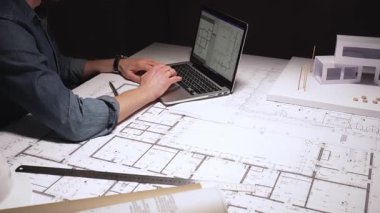 Mimar, pusula kullanarak büyük bina planlarına hassas ölçümler çiziyor, çağdaş bir ev tasarımı için mimari planlar üzerinde çalışıyor, bir dizüstü bilgisayar ve fiziksel model ile.