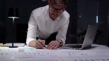 Geç saatlere kadar çalışan bir işadamı, kağıt belgelerindeki finansal veri ve grafikleri analiz ederken loş bir ofis ortamında dizüstü bilgisayar kullanarak, kendini adamışlık ve fazla mesai gösteriyor.