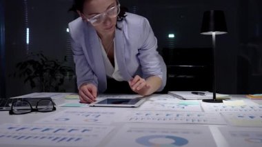 İş kadını masasında mali raporları ve çizelgeleri yoğun bir şekilde gözden geçiriyor, sunum hazırlamak ve bilgilendirilmiş yönetim kararları almak için notlar alıyor ve planlama stratejisi uyguluyor.