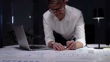 Geç saatlere kadar çalışan bir işadamı, kağıt belgelerindeki finansal veri ve grafikleri analiz ederken loş bir ofis ortamında dizüstü bilgisayar kullanarak, kendini adamışlık ve fazla mesai gösteriyor.