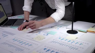 Meşgul profesyonel yönetici, çok sayıda finansal raporu ve iş belgelerini aktif olarak analiz ederek, bir dizüstü bilgisayar ve kalemle istatistikleri gözden geçirmek için, parlak bir şekilde aydınlatılmış bir masa üzerinde çalışmaktadır.