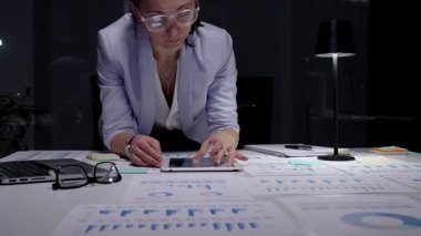 İş kadını masasında mali raporları ve çizelgeleri yoğun bir şekilde gözden geçiriyor, sunum hazırlamak ve bilgilendirilmiş yönetim kararları almak için notlar alıyor ve planlama stratejisi uyguluyor.