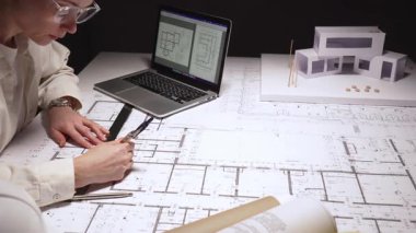Modern konut projesini tasarlayan mimar, cetvel ve kalemle planlar çizen, fiziksel bir modelle yapıyı görselleştiren, teknik planlar ve geliştirme için dizüstü bilgisayar kullanan