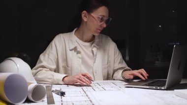 Güvenlik gözlüğü takan odaklanmış kadın mimar, iş yerindeki mimari planlara kesin çizgiler çizerek dizüstü bilgisayar ve beyaz şapka dışında modern bina planları oluşturuyor.