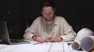 Güvenlik gözlüğü takan odaklanmış kadın mimar, iş yerindeki mimari planlara kesin çizgiler çizerek dizüstü bilgisayar ve beyaz şapka dışında modern bina planları oluşturuyor.