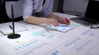 Finansal tablolar ve iş raporlarını incelemek için tablet kullanan profesyonel bir iş kadını strateji ve planlama için veri belgeleriyle dolu bir ofis masasında geç saatlere kadar çalışıyor.