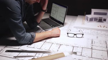 Mimar, pusula kullanarak büyük bina planlarına hassas ölçümler çiziyor, çağdaş bir ev tasarımı için mimari planlar üzerinde çalışıyor, bir dizüstü bilgisayar ve fiziksel model ile.