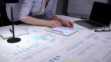Finansal tablolar ve iş raporlarını incelemek için tablet kullanan profesyonel bir iş kadını strateji ve planlama için veri belgeleriyle dolu bir ofis masasında geç saatlere kadar çalışıyor.
