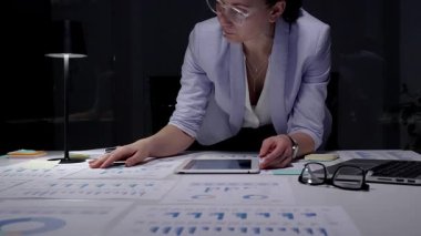 İş kadını masasında mali raporları ve çizelgeleri yoğun bir şekilde gözden geçiriyor, sunum hazırlamak ve bilgilendirilmiş yönetim kararları almak için notlar alıyor ve planlama stratejisi uyguluyor.