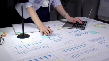 Ekonomik raporları ve hesap tablolarını bir masada inceleyen, stratejik planlama ve yatırım kararları için notlar alırken çizelgeleri ve dizüstü bilgisayarların grafiklerini analiz eden iş adamı.