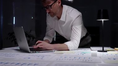 Geç saatlere kadar çalışan bir işadamı, kağıt belgelerindeki finansal veri ve grafikleri analiz ederken loş bir ofis ortamında dizüstü bilgisayar kullanarak, kendini adamışlık ve fazla mesai gösteriyor.