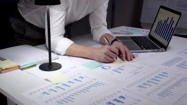 İş adamı finansal verileri ve istatistikleri analiz ediyor, ekranda grafikler ve çizelgeler olan bir dizüstü bilgisayarda yazıyor ve loş bir ofiste masaya yayılmış raporları inceliyor.
