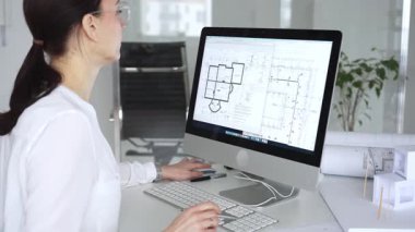 Mimari planlar ve bina modelleriyle çevrili bir masada, bir masaüstü bilgisayarı ve klavyeyi kullanarak gözlük takan mimar. İş ve mimari kavramı