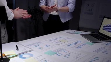 Karanlık bir ofiste el çırpan iş adamları, veri tabloları, finansal grafikler ve çalışan dizüstü bilgisayarlarla çevrili bir gece geç saatlerde yapılan toplantıda elde edilen başarıları kabul ediyorlar.