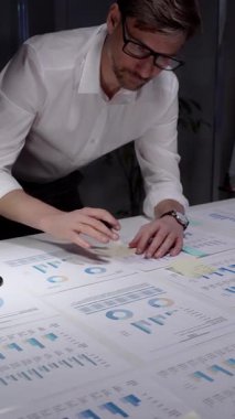 Profesyonel iş adamı masaya yaslanıyor. Gece geç saatlerde finansal veri ve çizelgeleri analiz ediyorum. Loş bir ofis ortamında fazla mesai yapmak. Ayrıntılı raporlara ve stratejik planlamaya odaklanmış.