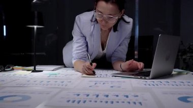 Finansal raporlar ve çizelgelerle kaplı bir masada duran bir kadın masa lambasının yumuşak parıltısı altında özenle bir dizüstü bilgisayar üzerinde çalışıyor, kendini işine adamış ve iş verilerinin detaylı analizini yapıyor.