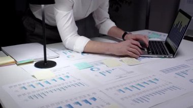 İş adamı finansal verileri ve istatistikleri analiz ediyor, ekranda grafikler ve çizelgeler olan bir dizüstü bilgisayarda yazıyor ve loş bir ofiste masaya yayılmış raporları inceliyor.