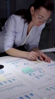 İş kadını masasında mali raporları ve çizelgeleri yoğun bir şekilde gözden geçiriyor, sunum hazırlamak ve bilgilendirilmiş yönetim kararları almak için notlar alıyor ve planlama stratejisi uyguluyor.