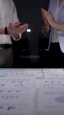 Karanlık bir ofiste el çırpan iş adamları, veri tabloları, finansal grafikler ve çalışan dizüstü bilgisayarlarla çevrili bir gece geç saatlerde yapılan toplantıda elde edilen başarıları kabul ediyorlar.