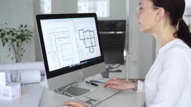 Mimari planlar ve bina modelleriyle çevrili bir masada, bir masaüstü bilgisayarı ve klavyeyi kullanarak gözlük takan mimar. İş ve mimari kavramı