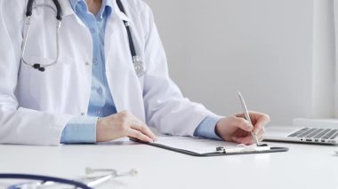 Sağlık evraklarını tamamlayan kadın doktor, dizüstü bilgisayarın yanına yerleştirilmiş bir pano ve cam kenarında iyi aydınlatılmış bir klinik çalışma alanında stetoskop. İlaç ve sağlık hizmetleri kavramı.