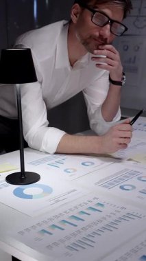 Profesyonel iş adamı masaya yaslanıyor. Gece geç saatlerde finansal veri ve çizelgeleri analiz ediyorum. Loş bir ofis ortamında fazla mesai yapmak. Ayrıntılı raporlara ve stratejik planlamaya odaklanmış.