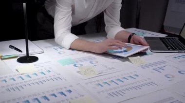 Meşgul profesyonel yönetici, çok sayıda finansal raporu ve iş belgelerini aktif olarak analiz ederek, bir dizüstü bilgisayar ve kalemle istatistikleri gözden geçirmek için, parlak bir şekilde aydınlatılmış bir masa üzerinde çalışmaktadır.