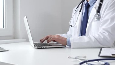 Doktor özenle bir dizüstü bilgisayarda yazıyor, muhtemelen hasta kayıtlarını güncelliyor veya araştırma yapıyor, modern bir tıbbi ortamda sağlık ve teknolojinin kesişimini gösteriyor.