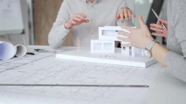 Bir konut projesinde işbirliği yapan mimarlar, planlar ve ölçekli bir modeli bir masada inceleyerek, tasarım detaylarını, inşaat planlarını ve gelecekteki gelişimi tartışarak,