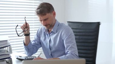 Mavi gömlekli ve gözlüklü profesyonel bir muhasebeci ofis ortamında mali raporları incelerken fatura hesaplıyor ve faturaları yönetiyor. İş dünyasında denetim ve vergi teması.